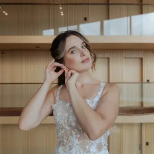 Bruid in kanten trouwjurk doet haar oorbel in tijdens getting ready moment in modern gebouw met houten interieur.